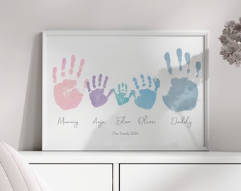 May include: A white framed print with five handprints in different colors, pink, purple, blue, light blue, and blue. The handprints are arranged in a line from left to right. Below the handprints are the names "Mummy", "Anja", "Eden", "Oliver", and "Daddy". The text "Our Family 2022" is below the names.