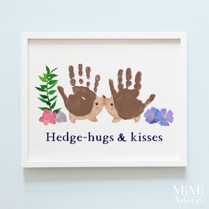 Op de afbeelding: Een wit ingelijst print met een handafdruk-kunstontwerp van twee egels die elkaar kussen. De egels zijn bruin en hebben schattige gezichtjes. De print heeft groene bladeren, roze bloemen en blauwe bloemen. De tekst "Hedge-hugs & kisses" is in donkerblauw onder de egels gedrukt.