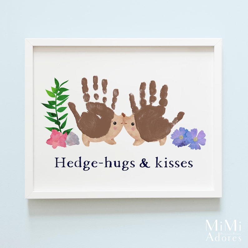 Op de afbeelding: Een wit ingelijst print met een handafdruk-kunstontwerp van twee egels die elkaar kussen. De egels zijn bruin en hebben schattige gezichtjes. De print heeft groene bladeren, roze bloemen en blauwe bloemen. De tekst "Hedge-hugs & kisses" is in donkerblauw onder de egels gedrukt.