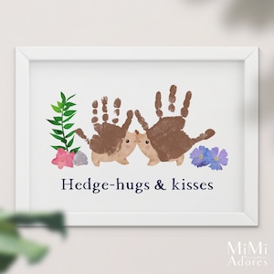 Op de afbeelding: Een ingelijst print met een handafdruk-kunstontwerp van twee egels die elkaar omhelzen. De egels zijn bruin en hebben schattige gezichtjes. De print heeft de tekst "Hedge-hugs & kisses" in donkerblauwe letters.