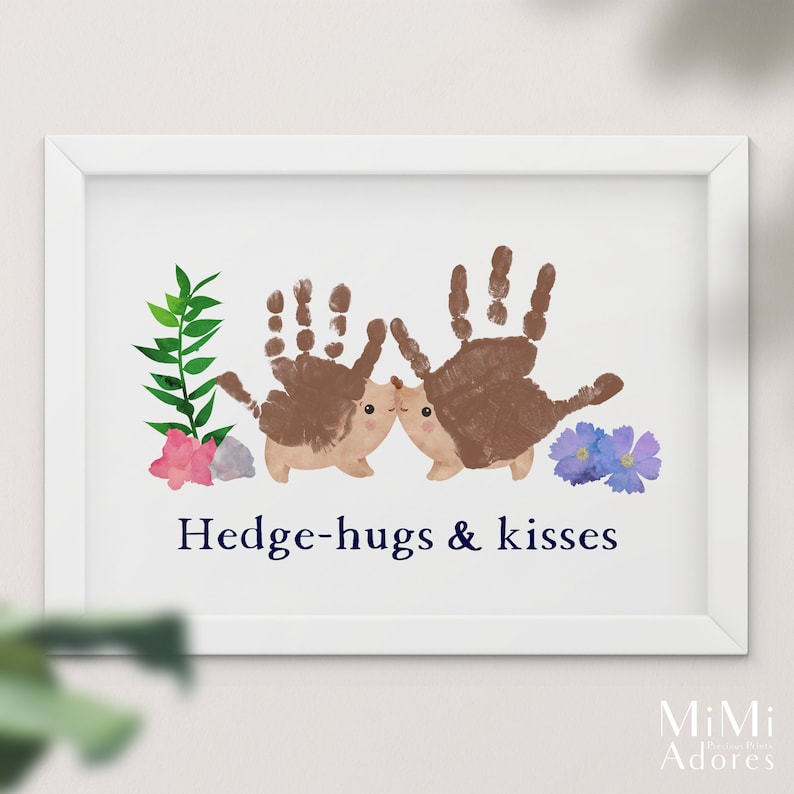 Op de afbeelding: Een ingelijst print met een handafdruk-kunstontwerp van twee egels die elkaar omhelzen. De egels zijn bruin en hebben schattige gezichtjes. De print heeft de tekst "Hedge-hugs & kisses" in donkerblauwe letters.