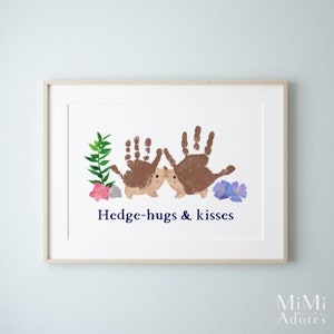 Op de afbeelding: Een ingelijst print met een handafdruk-kunstontwerp van twee egels die elkaar kussen. De egels zijn bruin en hebben schattige gezichtjes. De print heeft een witte achtergrond met groene bladeren en paarse bloemen. De tekst "Hedge-hugs & kisses" is in blauw onder de egels gedrukt.