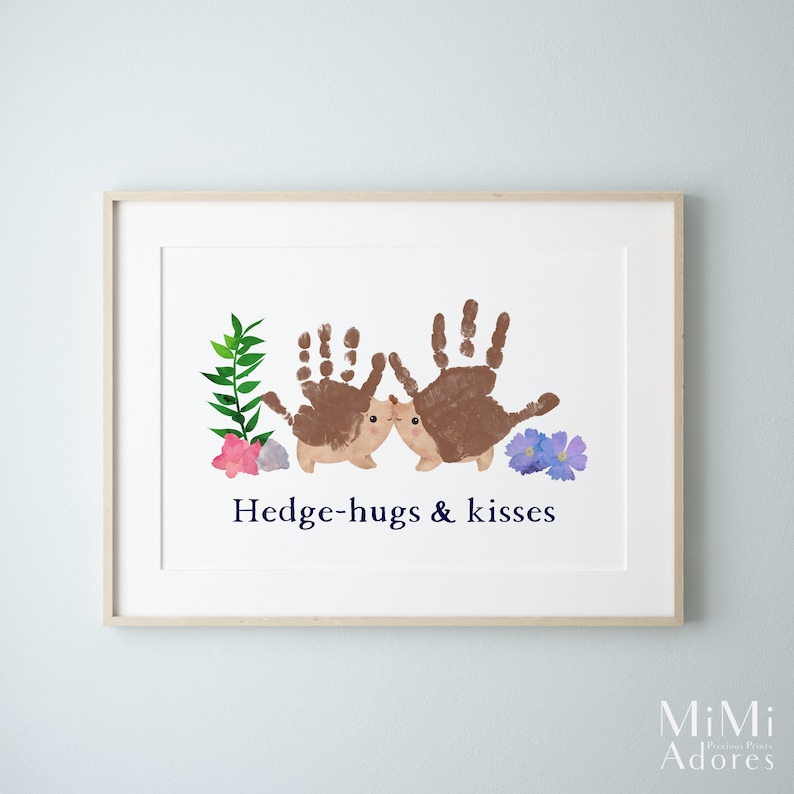 Op de afbeelding: Een ingelijst print met een handafdruk-kunstontwerp van twee egels die elkaar kussen. De egels zijn bruin en hebben schattige gezichtjes. De print heeft een witte achtergrond met groene bladeren en paarse bloemen. De tekst "Hedge-hugs & kisses" is in blauw onder de egels gedrukt.