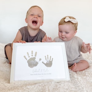 Könnte beinhalten: Ein weißer gerahmter Druck mit zwei silbernen Handabdrücken und dem Text "Gabriel & Psalm have hands down the best Daddy! June 2023". Zwei Kinder, ein Junge und ein Mädchen, halten den Druck.