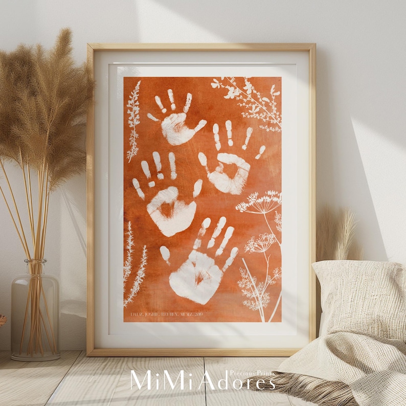 May include: A framed print with a rust-colored background featuring white handprints and white silhouettes of plants. The text "Our Family, Their Hands, Our Memories" is printed at the bottom of the print.