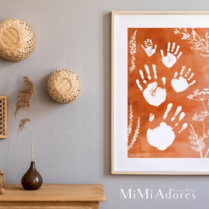 May include: A framed print with a watercolor background in shades of orange and brown. White handprints are layered on top of the background. The print includes the text "Our Family 2024" and the logo "MiMi Adore's Precious Prints".