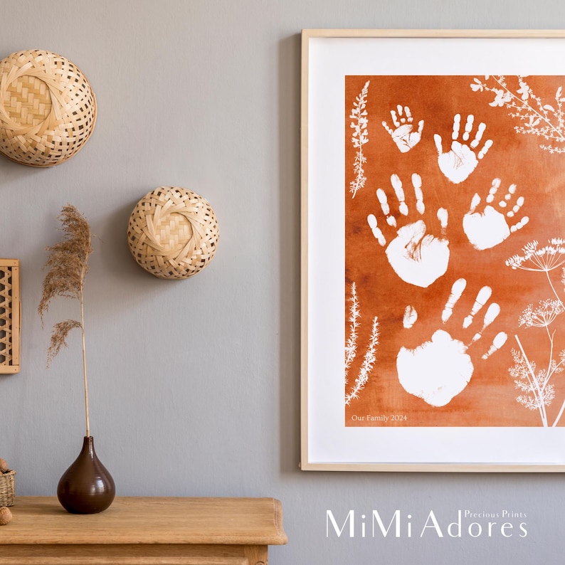 May include: A framed print with a watercolor background in shades of orange and brown. White handprints are layered on top of the background. The print includes the text "Our Family 2024" and the logo "MiMi Adore's Precious Prints".