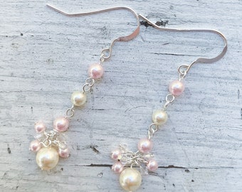May include: Elegant dangle earrings featuring a cluster of cream and pink faux pearls. The earrings have a silver-tone hook and chain design, with a larger pearl at the bottom of the cluster. A delicate and feminine accessory.