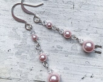May include: A pair of dangle earrings featuring pink faux pearls and clear crystal beads. The earrings have silver-colored fish hook ear wires and are connected by small silver-colored rings. The pearls vary in size, with the largest at the bottom.