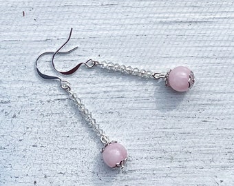 May include: Dangle earrings featuring a light pink, round bead with a silver-colored decorative cap. The beads are suspended from a silver-colored hook with a strand of small, clear beads.