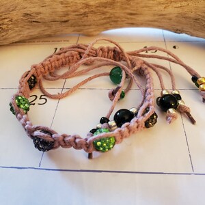 May include: A pink macrame bracelet with green and black beads. The bracelet has a tassel on each end.