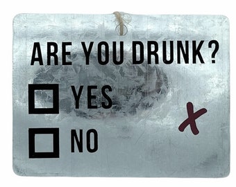 May include: A rectangular metal sign with the text "ARE YOU DRUNK?" in black. Below are two boxes with "YES" and "NO" options. A red "X" marks the "YES" box. A string is attached at the top.