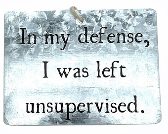 May include: A rectangular metal sign with the text "In my defense, I was left unsupervised." The sign has a textured, galvanized metal appearance and a natural twine loop for hanging. The text is in a simple, sans-serif font.
