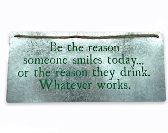 May include: A rectangular metal sign with the text "Be the reason someone smiles today... or the reason they drink. Whatever works." in green. The sign has a rustic, galvanized finish and a twine hanger.