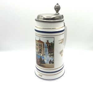 May include: White ceramic beer stein with a silver lid and handle. The stein features a colorful image of a building and people. Gold and blue bands encircle the stein, with the year "1995" at the base. A decorative piece for collectors.