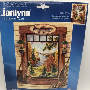 May include: Blue and white packaging for a counted cross stitch kit depicting a window scene with a lake, trees, and a trout. The kit is titled "Sportsman's Dream" and includes 14-count Aida fabric, 6-strand cotton floss, needle, graph, floss cards, and instructions. The kit is designed by Nancy Rossi.