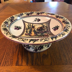 Op de afbeelding: Een keramische voetkom met een crèmekleurige basis en een decoratieve rand. De kom heeft een ontwerp met twee konijnen omringd door groenten en bloemen in tinten groen, bruin en paars.