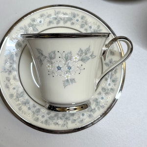 Könnte beinhalten: Eine weiße Teetasse und Untertasse mit silbernem Rand und Blumenmuster. Das Blumenmuster zeigt blaue und weiße Blumen mit grünen Blättern.