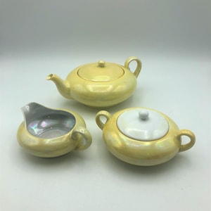 May include: A set of three yellow ceramic tea service pieces, including a teapot, creamer, and sugar bowl with a lid. The pieces have a pearlescent finish and are decorated with a subtle iridescent sheen.