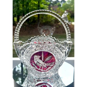 May include: A clear crystal basket with a handle and a bird figurine on top. The basket features a decorative pattern and a circular, magenta-colored emblem with a bird and foliage design. The basket is displayed on a reflective surface.