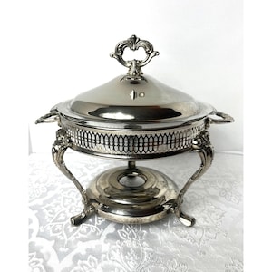May include: A silver-colored chafing dish with a lid and ornate handles. The dish has a decorative rim and sits on a stand with curved legs. The lid features a decorative handle.