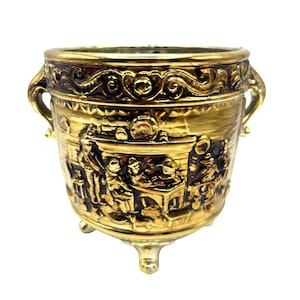 May include: A gold ceramic planter with a raised design of people gathered around a table. The planter has two handles and three feet.