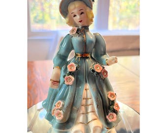 May include: A vintage porcelain figurine of a woman in a long, light blue dress adorned with pink roses. The figurine has blonde hair, a matching hat, and gold accents. The base is white and the figurine is on a mirrored surface.