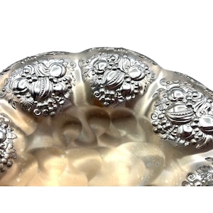 May include: A silver serving bowl with an ornate design of grapes and leaves. The bowl has a scalloped edge and a hammered finish.
