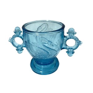 May include: A light blue glass sugar bowl with two swan handles. The bowl has a textured design and a pedestal base.