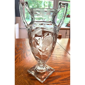 May include: A clear glass vase with two handles and a square base. The vase is etched with a floral design.
