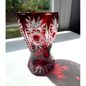 May include: A vibrant ruby red crystal vase, featuring detailed starburst and circular cut patterns. The vase has a flared top and a rounded base, creating a dramatic shadow. The cut crystal design reflects light, enhancing its decorative appeal.