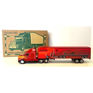 May include: A red and orange die-cast metal Freightliner cab with a matching trailer. The trailer features a BFGoodrich logo and the text "Now available in larger sizes". The box shows a green illustration of the truck.