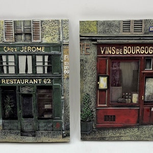 May include: Two vintage-style Parisian storefront plaques. The left plaque features a green storefront with the text "Chez Jerome" and "Restaurant 62". The right plaque features a red storefront with the text "Vins de Bourgogne".