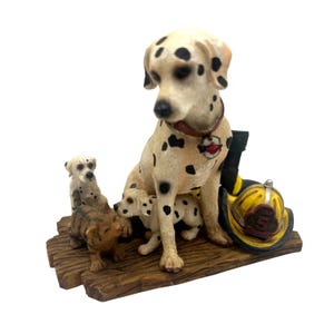 Puede incluir: Figura de un perro dálmata con manchas negras, sentado con tres cachorros y un pequeño gato marrón sobre una base de madera. Un casco de bombero amarillo y una bota negra están al lado.