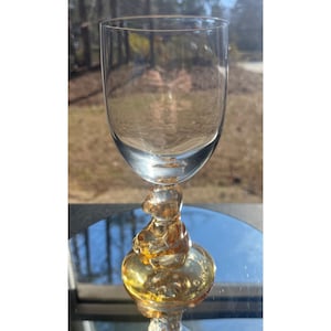 May include: A clear wine glass with a decorative amber-colored base featuring a bear figure. The glass has a rounded bowl and a slender stem, perfect for serving wine or other beverages. The glass is set against a blurred outdoor background.