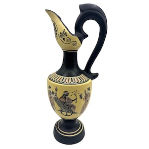 Puede incluir: Jarra de cerámica negra y beige con asa y pico curvos. La jarra presenta una escena pintada de figuras con atuendo clásico. La base y la parte superior son negras, mientras que el cuerpo es beige con diseños decorativos.
