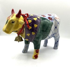 May include: A colorful ceramic cow figurine with a patchwork design, yellow, green, purple, blue, and white colors, with a red bow and a gold bell around its neck.