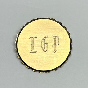 Puede incluir: Pin redondo de metal dorado con borde festoneado. El pin tiene las letras "LGP" grabadas en una fuente estilizada.