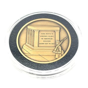 May include: A bronze coin with a soldier standing at attention in front of a tomb. The inscription reads "Here rests in honored glory an American soldier known but to God."