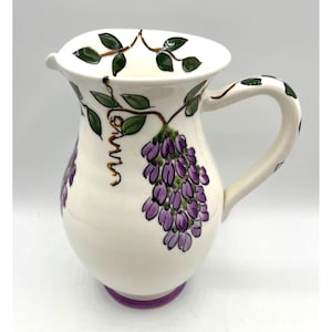 May include: White ceramic pitcher with a purple base and handle. The pitcher is decorated with purple wisteria flowers, green leaves, and brown vines. The handle and rim are also decorated with green leaves.