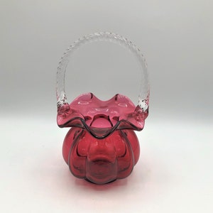May include: A small, ruby red glass basket with a clear glass handle. The basket has a ruffled rim and a rounded bottom.