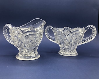 Op de afbeelding: Een vintage set van een roomkan en suikerpot van helder glas met ingewikkelde geslepen glasontwerpen. De stukken hebben een delicate, elegante uitstraling.