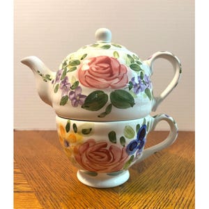 Peut inclure: Service à thé individuel en céramique blanche composé d'une théière et d'une tasse. L'ensemble est orné de roses roses, de fleurs violettes et de feuilles vertes peintes à la main. La tasse est sous la théière. Sur une surface en bois.