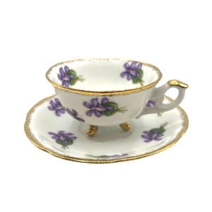 May include: A white teacup and saucer set with a gold trim and purple flower design. The teacup has three gold feet.