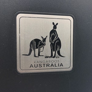 May include: A silver metal sign with two black silhouettes of kangaroos. The sign reads "Kangaroos Australia".