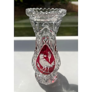 Könnte beinhalten: Eine klare Kristallvase mit einem roten und silbernen Vogeldesign. Die Vase hat ein rautenförmiges Muster und einen ausgestellten oberen Rand. Der Vogel ist mit einer Weizenähre dargestellt. Die Vase ist etwa 15 cm hoch.