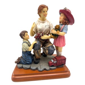 Puede incluir: Figura de un bombero con dos niños. El bombero, sentado, sostiene un camión de bomberos de juguete rojo. Un niño está arrodillado y una niña sostiene un oso de peluche. La base es de madera y las figuras están detalladas.