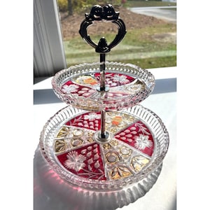 May include: Two-tiered glass serving tray with a decorative handle. The trays have a red and gold floral design. The edges are scalloped. The handle is silver-toned. This elegant serving piece is perfect for displaying desserts or appetizers.