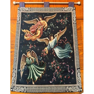 May include: A tapestry featuring three angels with golden wings, halos, and flowing robes in shades of pink, blue, and green. The angels are set against a dark blue background with roses and ribbons. The tapestry has a decorative border and is hung from a silver rod.