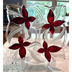 Può includere: Un piatto da portata in vetro trasparente di forma quadrata con quattro disegni di fiori rossi. Il piatto ha accenti bianchi e argentati ed è sostenuto da un supporto trasparente. Il design floreale aggiunge un tocco decorativo.